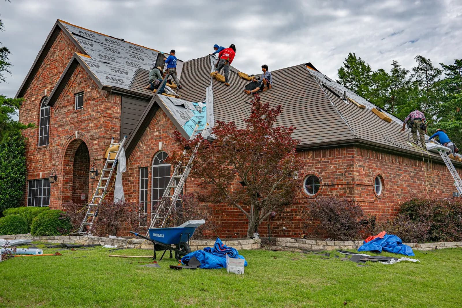 Crew of roofers working across the roof of a large brick home, with ladders, tarps and a wheelbarrow on the lawn below.