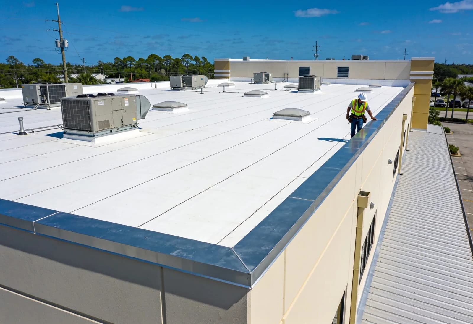 White flat membrane roof on a commercial building with rooftop HVAC units, skylights and metal edge trim, with a worker in a hard hat and safety vest walking the roof.
