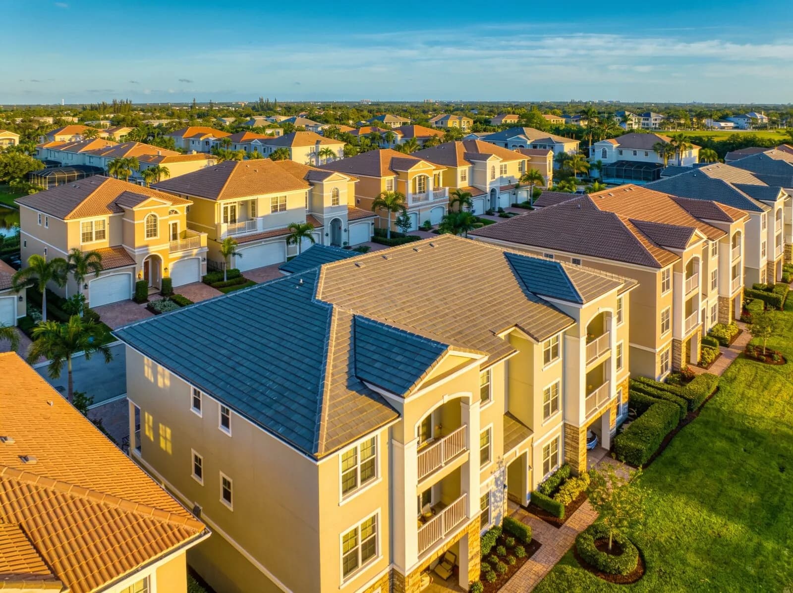 Aerial view of a Florida community of three-story yellow stucco townhome buildings with slate-blue and terracotta tile roofs, palm trees and green lawns in golden light.
