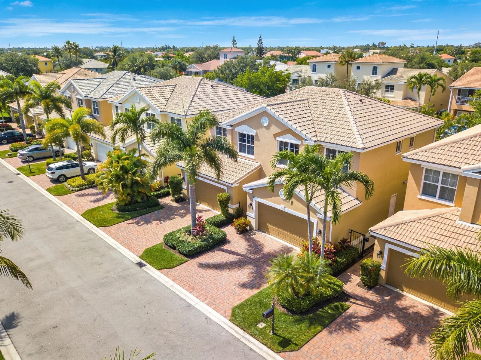 Aerial view of a row of two-story yellow stucco townhomes with beige tile roofs, palm trees and brick-paver driveways along a residential street in Florida.