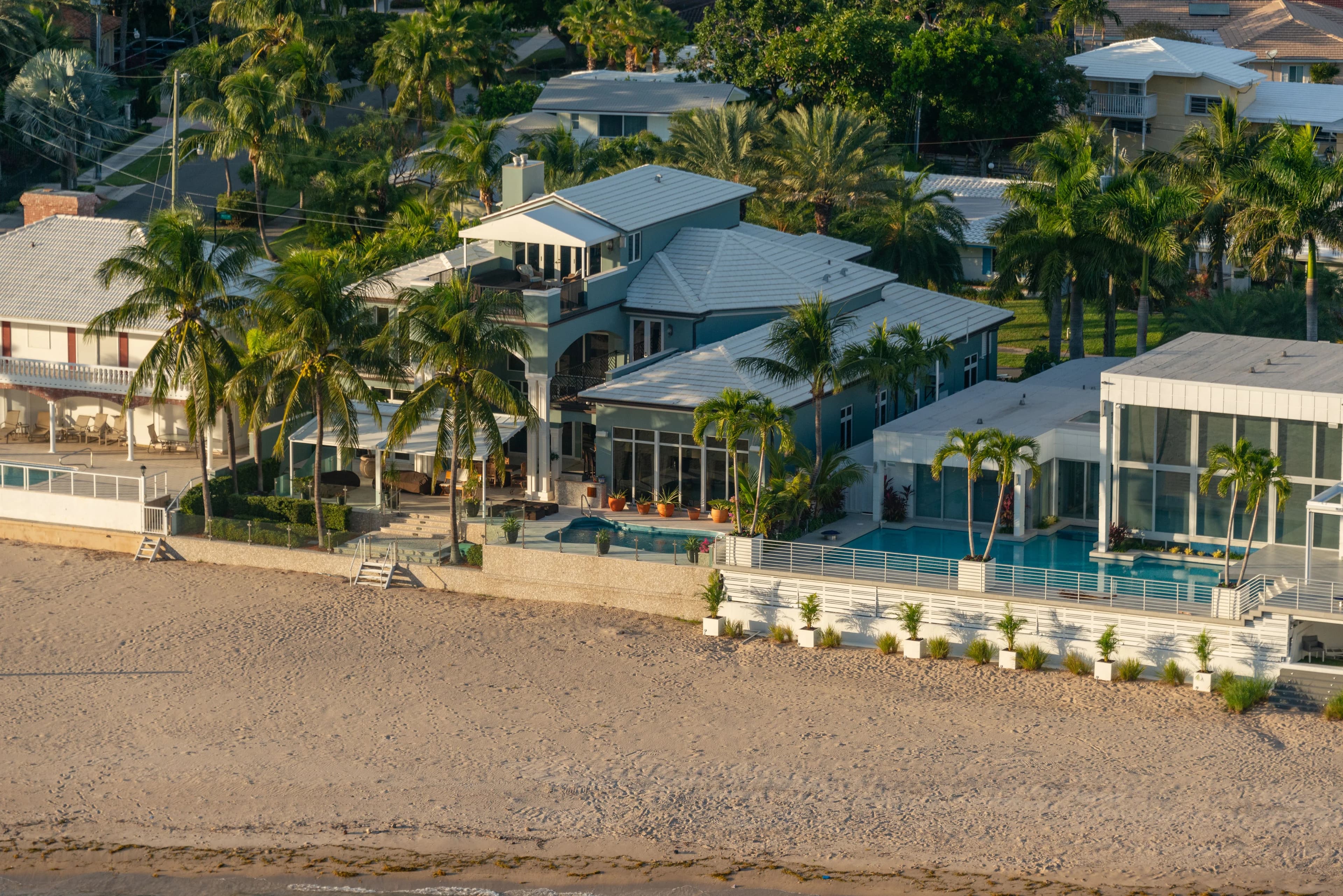 Aerial view of luxury beachfront homes with light-colored roofs, palm trees and pools in warm morning light.