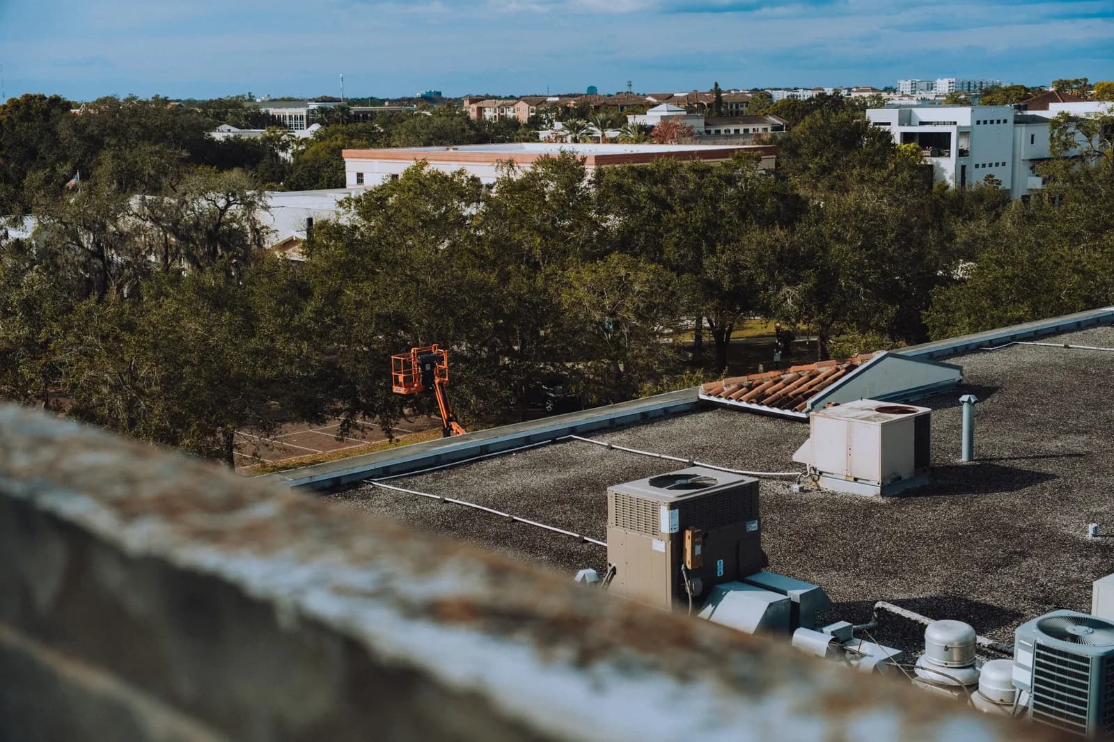 Low-slope commercial rooftop with packaged HVAC units and vents, seen past a parapet wall with oak trees and low buildings beyond.