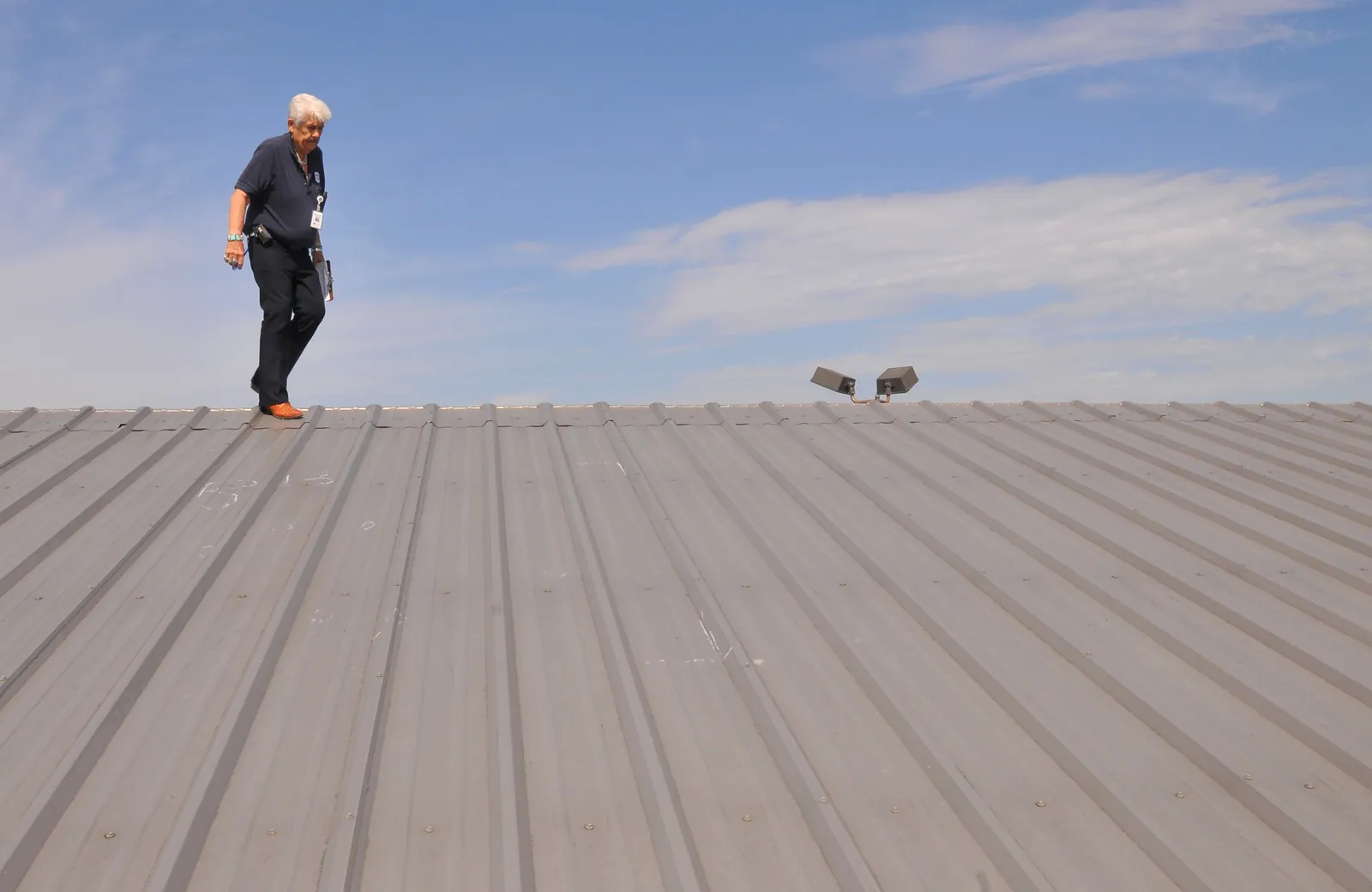Inspector walking along the ridge of a ribbed metal roof, checking the panels under a blue sky.
