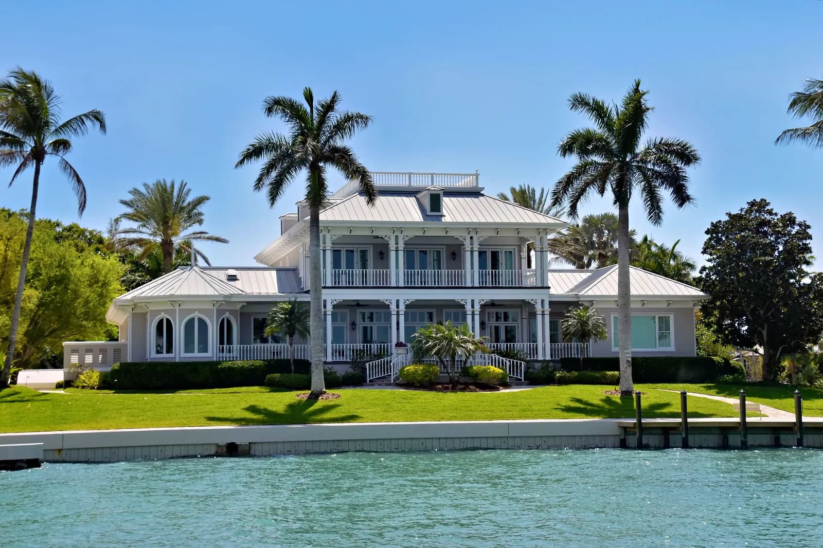 Two-story waterfront villa with a silver standing-seam metal roof, white wraparound porches and royal palms beside a seawall.