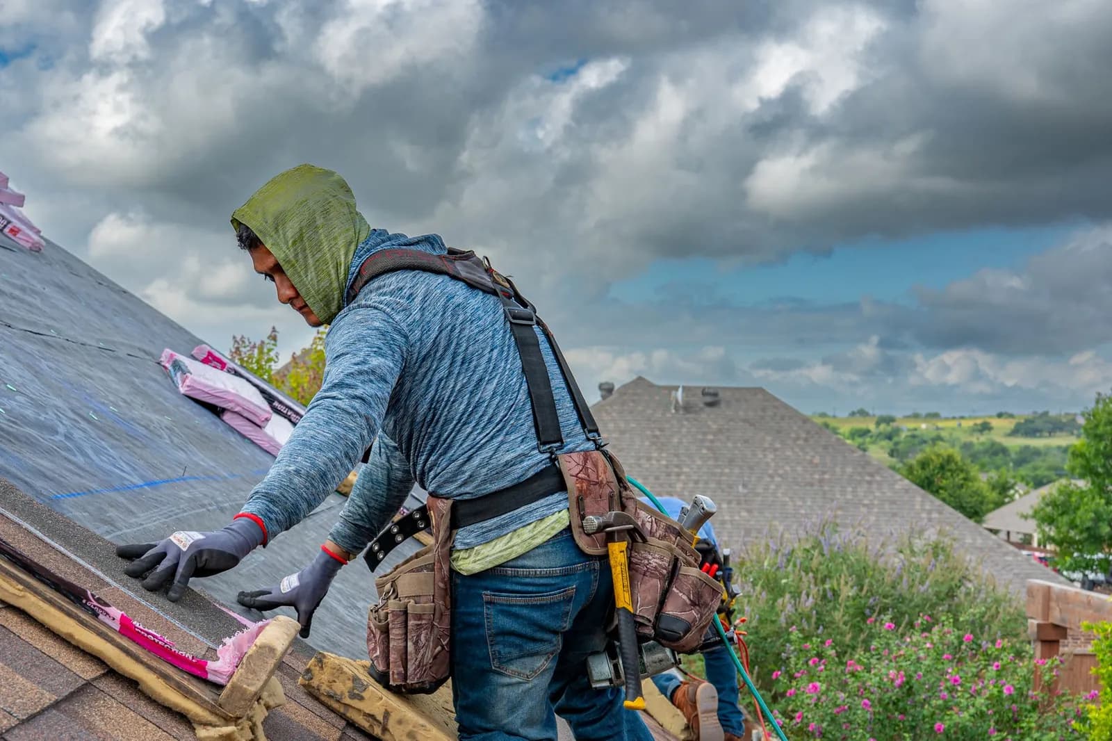 Roofer wearing gloves, a tool belt and a safety harness laying shingles along a roof edge under a cloudy sky.