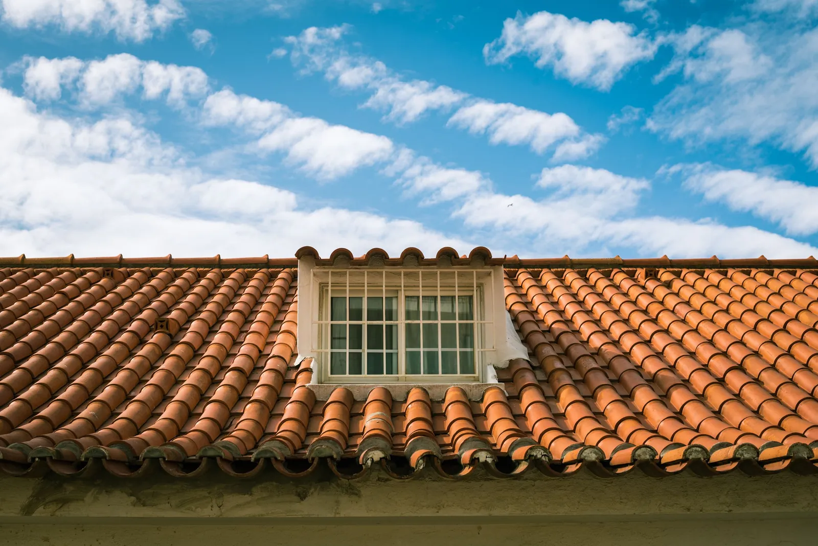 Close view of a terracotta barrel tile roof with a white dormer window under a blue sky streaked with clouds.